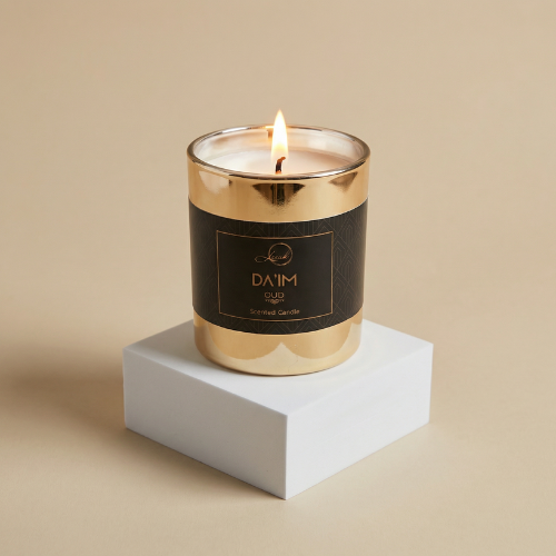 A lit DA'IM Oud scented candle by Leesh. The candle is housed in a reflective, metallic gold glass jar with a thick, matte black label wrapping around the center. The label features a subtle geometric pattern and the brand name "Leesh" and "DA'IM OUD" in elegant gold lettering. The candle sits on a solid white square pedestal against a warm beige background.