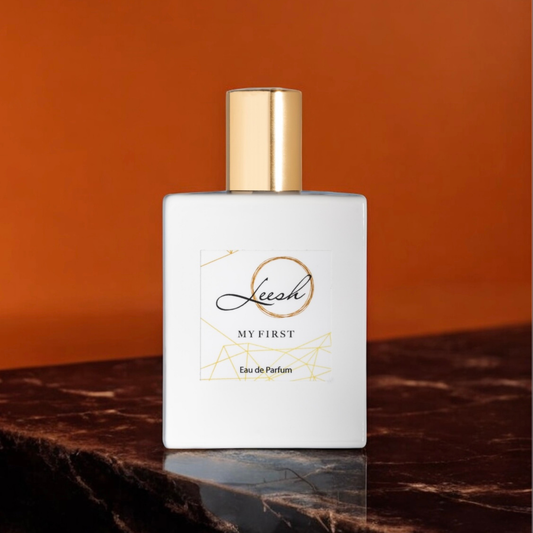 Her My First by Leesh — a flirty and empowering fragrance for women. Featuring patchouli, cedarwood, vanilla, jasmine, rose, ylang-ylang, amber, and musk. Elegant bottle shot with warm amber lighting, reflecting confidence and unforgettable moments.