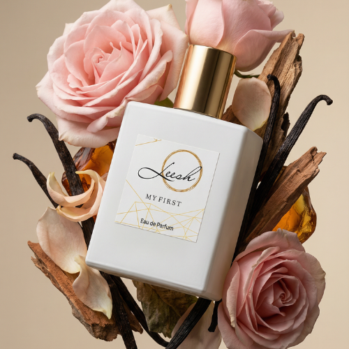 A dynamic product shot of 'Her My First' Eau de Parfum by Leesh Fragrances. The signature opaque white square bottle with a gold cap and geometric label is floating among its key fragrance notes. It is elegantly framed by blooming light pink roses, dark vanilla bean pods, textured wood pieces, and glowing amber resin, all set against a warm, soft beige background.