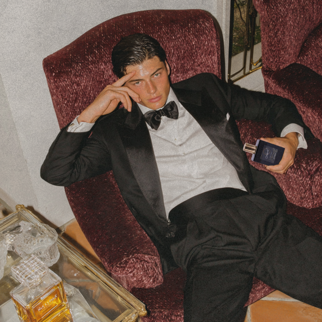 Man in a black tuxedo sitting on a red velvet chair with a bottle of perfume on a table. Model holding his first perfume by leesh fragrances at a new year 2026 party, styled in cinematic editorial luxury setting with warm lighting and shadows