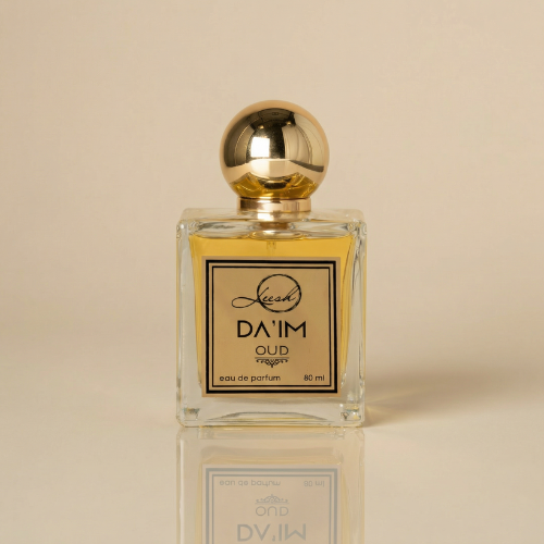 A clear, square glass perfume bottle of DA'IM Oud Eau de Parfum by Leesh, filled with a warm golden liquid. The bottle features a highly reflective, spherical gold cap and a metallic gold label with a black border. The brand and fragrance name are printed in elegant black typography. The bottle sits on a glossy beige surface, creating a sharp, mirrored reflection beneath it against a soft beige background.
