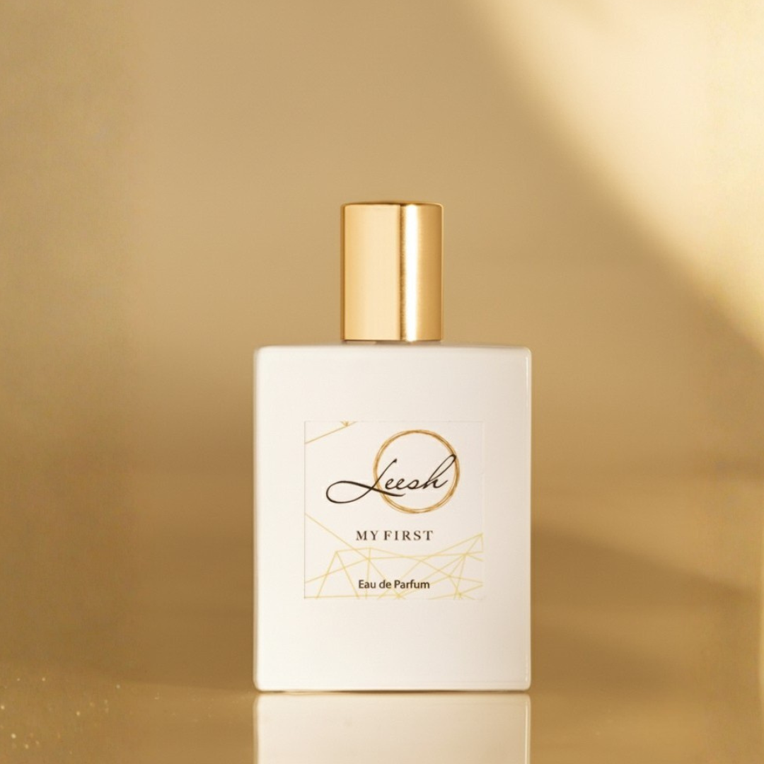 Perfume bottle labeled 'My First' with gold cap on a beige background - Her My First by Leesh — a flirty and empowering fragrance for women. Featuring patchouli, cedarwood, vanilla, jasmine, rose, ylang-ylang, amber, and musk. Elegant bottle shot with warm amber lighting, reflecting confidence and unforgettable moments.
