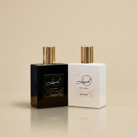 A side-by-side product shot of the 'My First' fragrance duo by UK brand Leesh Fragrances. On the left is 'His My First' in an opaque black square bottle with a black and gold geometric label. On the right is 'Her My First' in an opaque white square bottle with a matching white and gold label. Both feature metallic gold caps and cast clear reflections on a smooth, warm beige surface.