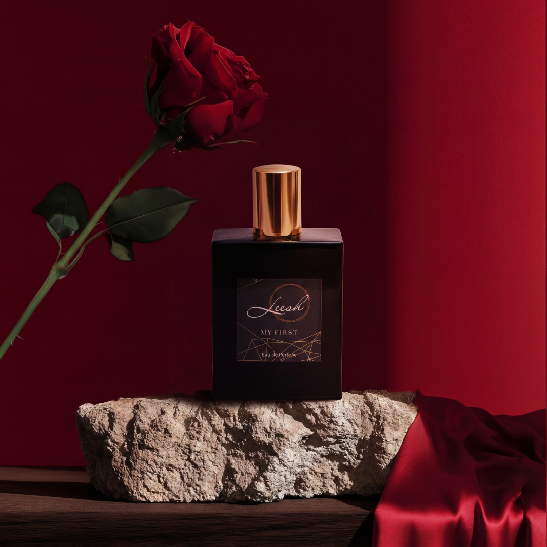 Perfume bottle with gold cap and red rose on a stone surface against a red background. His My First fragrance by Leesh Fragrances displayed on a wooden table with a red background and a rose, showcasing the Valentine’s Day scent notes of bergamot, black pepper, cedarwood and amber. 