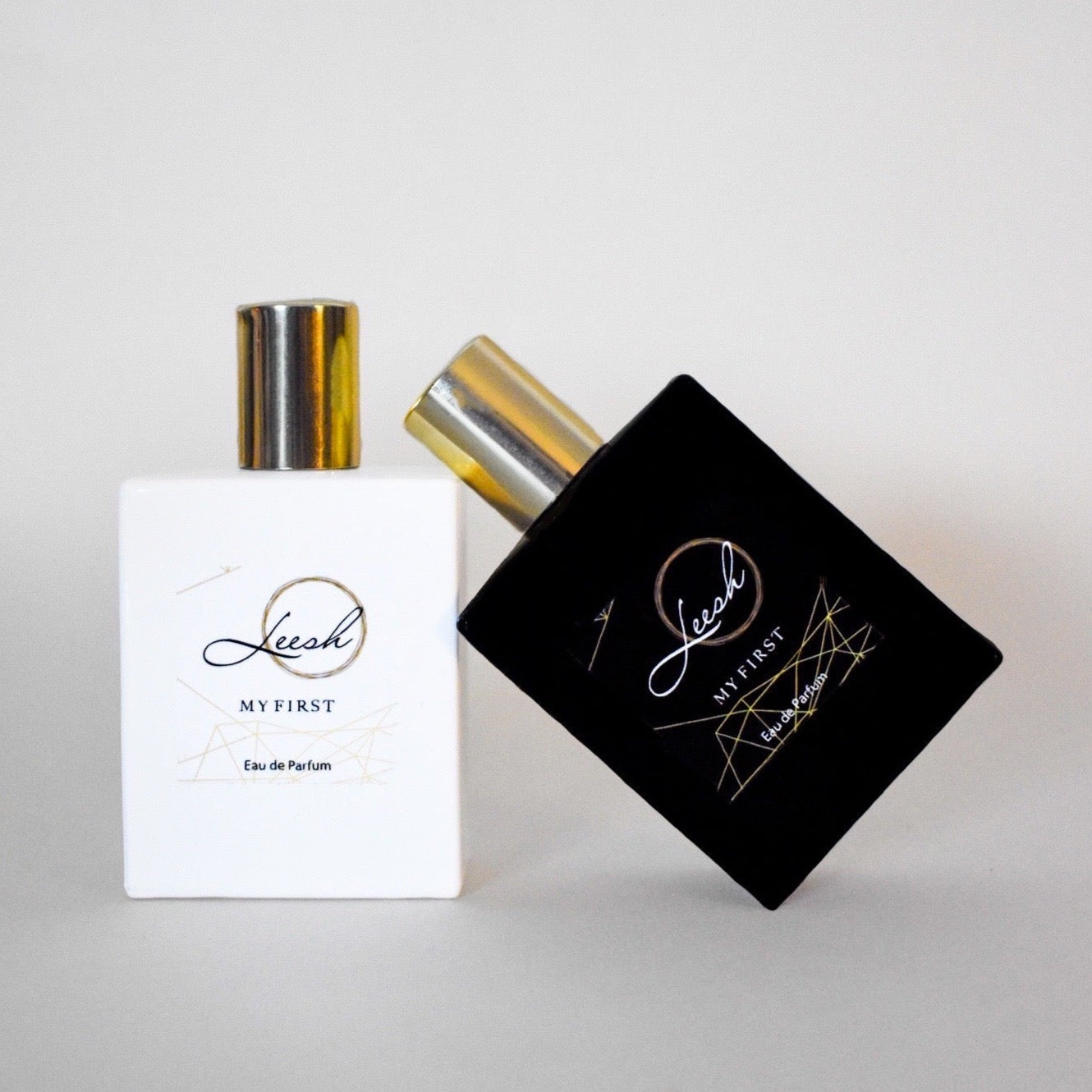 Shop Leesh Fragrances' luxurious Her First and His First fragrance bottles on sleek grey background. The elegant white bottle of Her First features a gleaming gold lid, while the sophisticated black bottle of His First, with matching gold lid, leans on Her First.